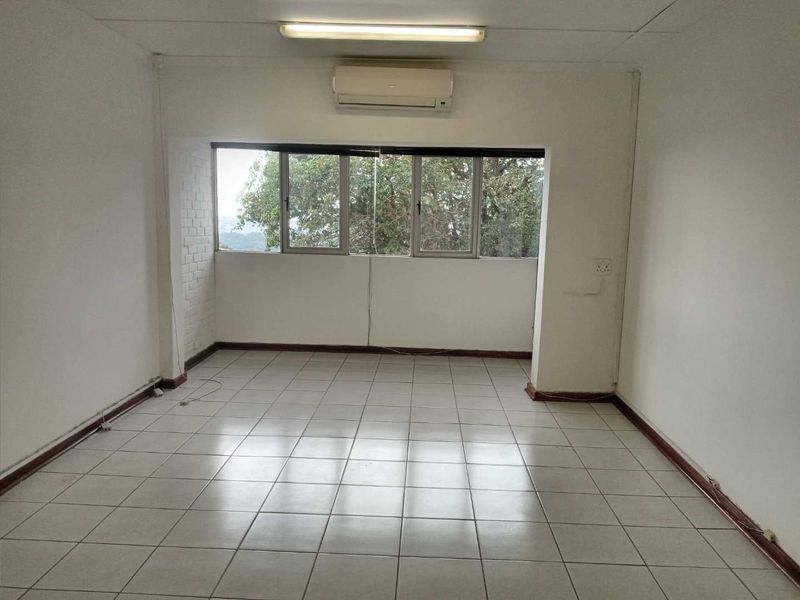 An empty office space featuring tiled flooring, a wall-mounted air conditioning unit, and large windows providing natural light and exterior views.