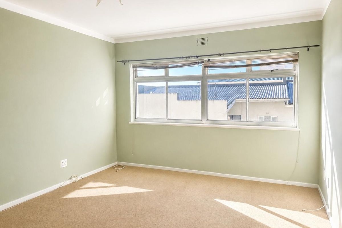 A bright, empty bedroom featuring neutral carpet flooring, soft green walls, and a large sliding window with a curtain rod.