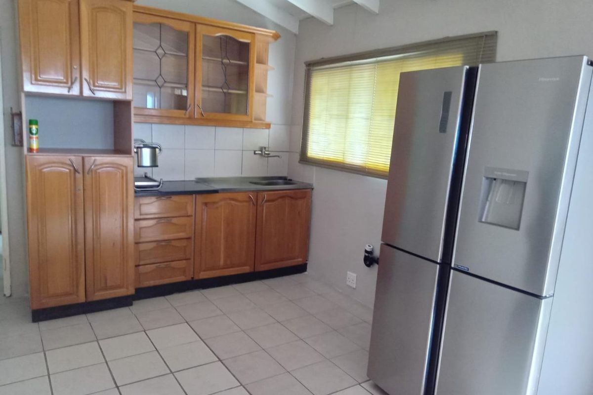 A functional kitchen featuring timber cabinetry, tiled splashbacks, and tiled flooring, complemented by exposed rafters and a window with fitted blinds.