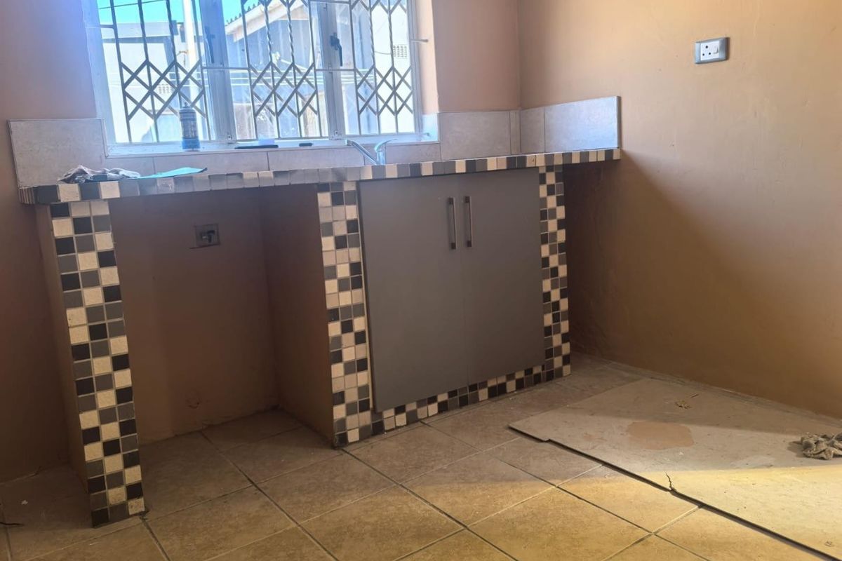 A basic kitchen area featuring a tiled countertop, built-in under-counter cabinetry, and a security gate over the window.