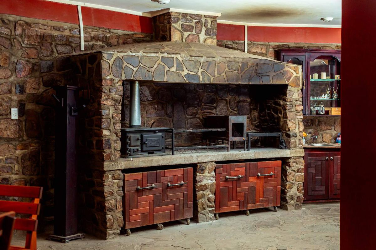 Rustic indoor braai area featuring a large stone-clad fireplace, built-in wood-burning stove, and custom wooden cabinetry in a farm-style setting.