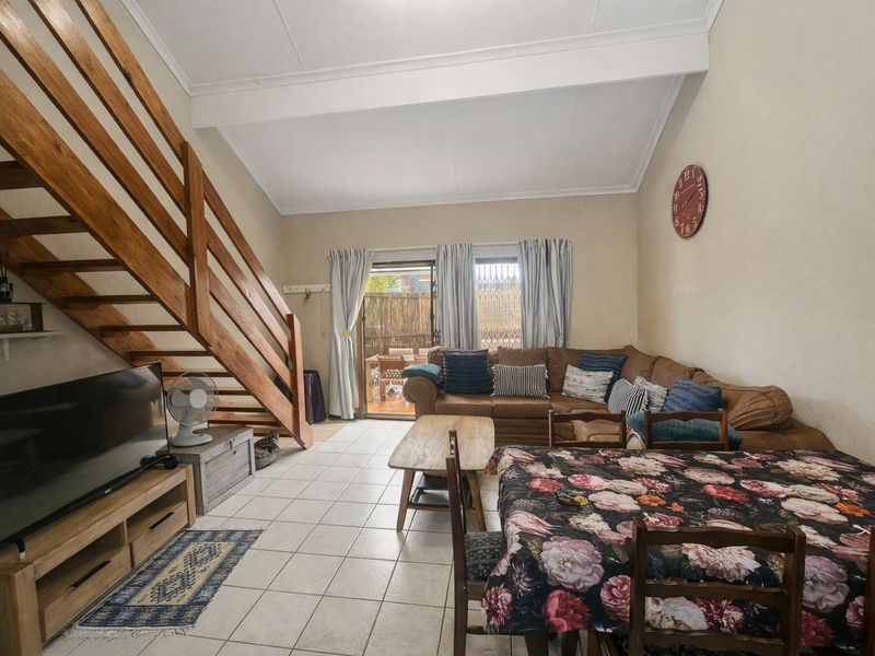 Open-plan living and dining area featuring tiled floors, a vaulted ceiling, and a wooden staircase leading to the upper level with patio access.