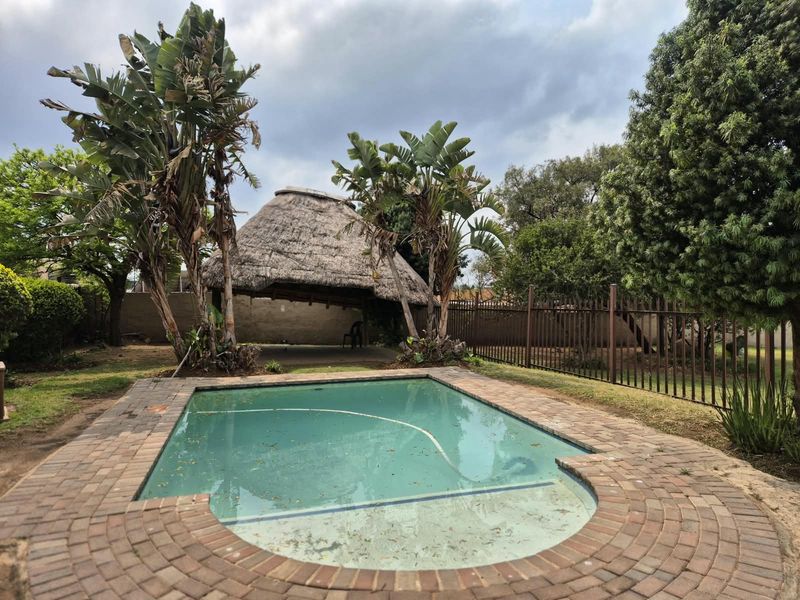 Outdoor pool area featuring a swimming pool with brick coping and a traditional thatch lapa structure set within a fenced garden.