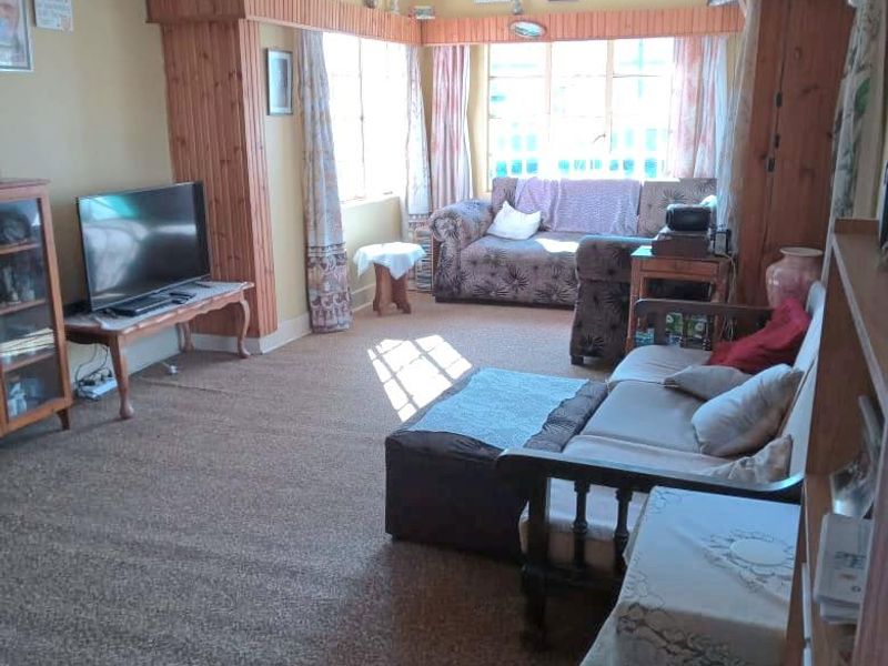 A living room featuring dated timber wall paneling and ceiling beams, with large windows providing natural light and carpeted flooring throughout.