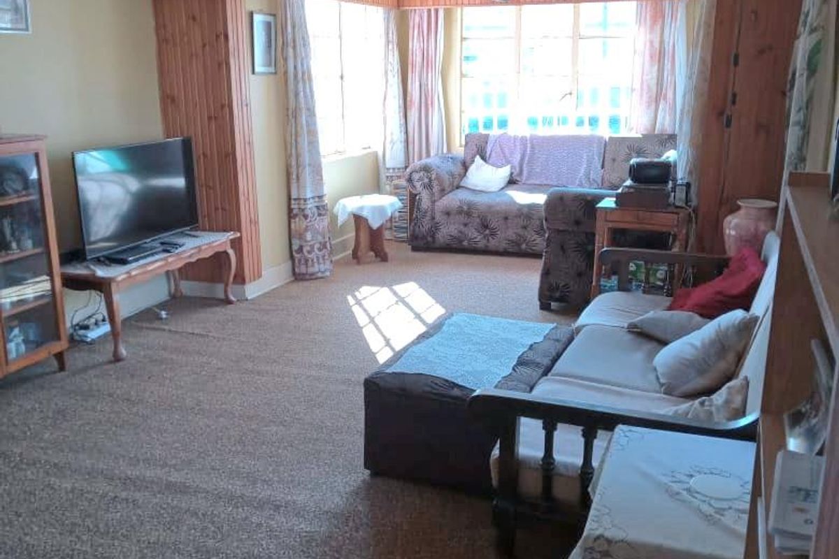 A living room featuring dated timber wall paneling and ceiling beams, with large windows providing natural light and carpeted flooring throughout.