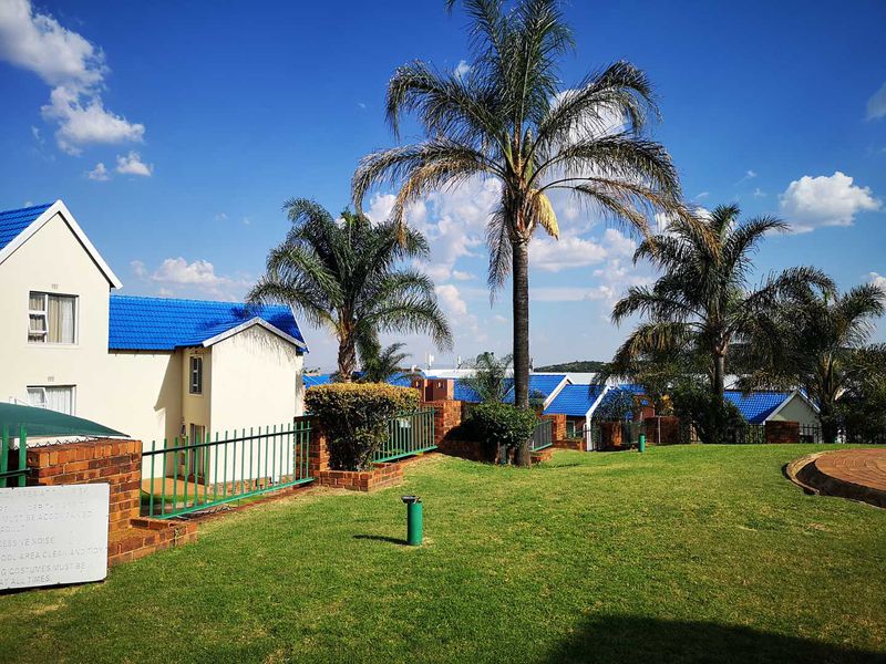 A well-maintained communal garden area featuring manicured lawns, mature palm trees, and brick boundary walls with metal fencing surrounding the townhouse complex.