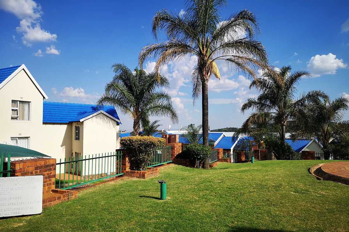 A well-maintained communal garden area featuring manicured lawns, mature palm trees, and brick boundary walls with metal fencing surrounding the townhouse complex.
