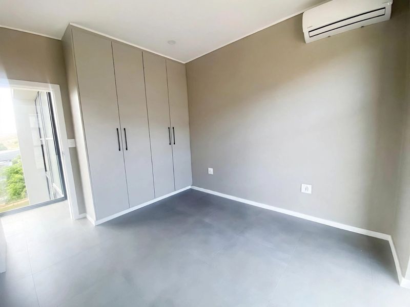 A clean, modern bedroom featuring neutral-toned built-in cupboards, tiled flooring, and a wall-mounted air conditioning unit with access to an outdoor area.