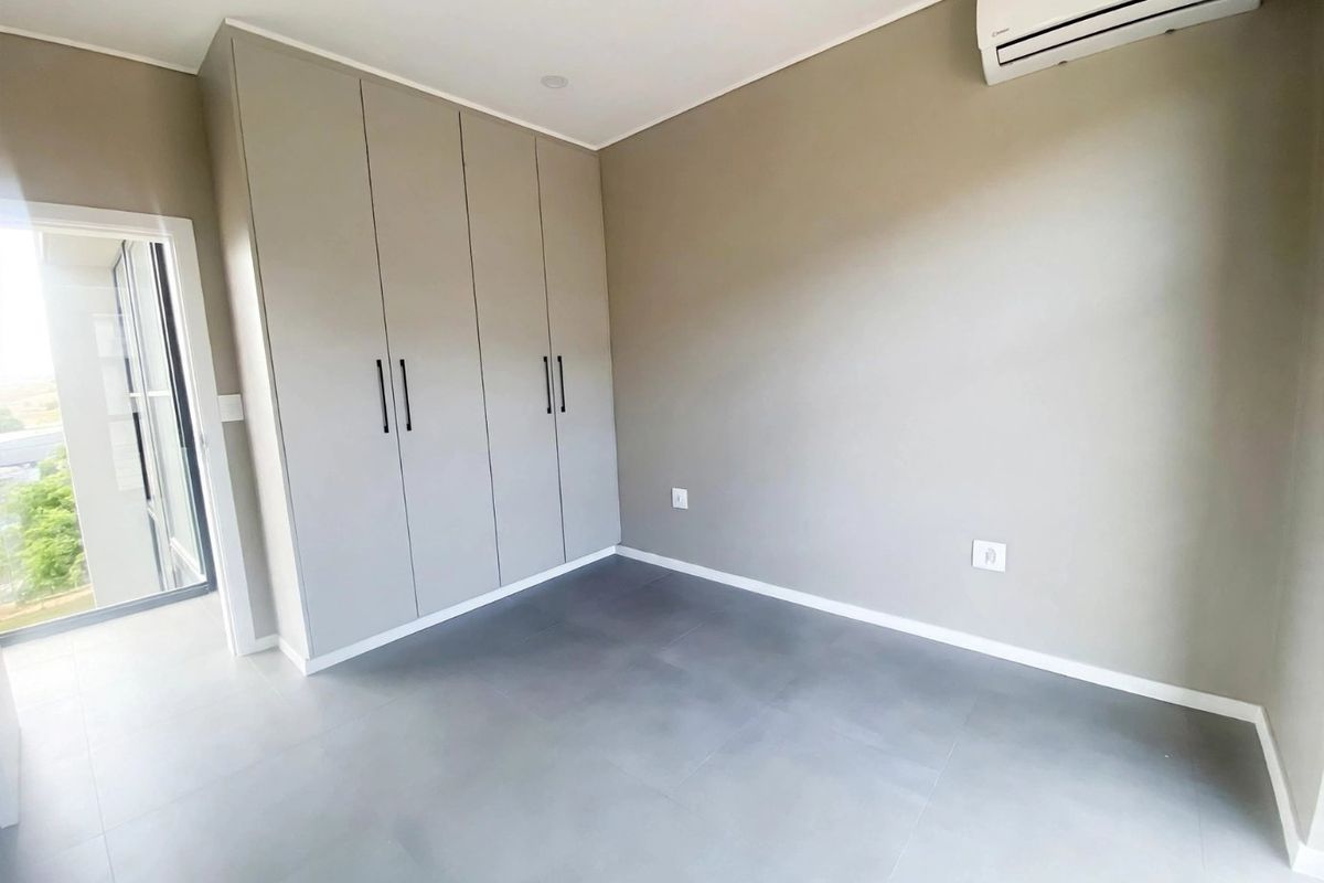 A clean, modern bedroom featuring neutral-toned built-in cupboards, tiled flooring, and a wall-mounted air conditioning unit with access to an outdoor area.