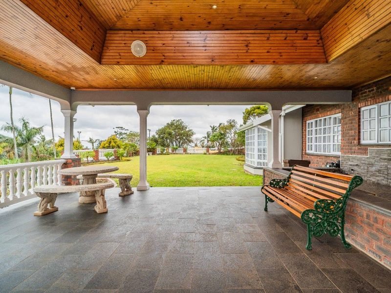 A spacious covered patio featuring a timber-clad ceiling, stone tiled flooring, and decorative columns, overlooking a well-maintained garden.