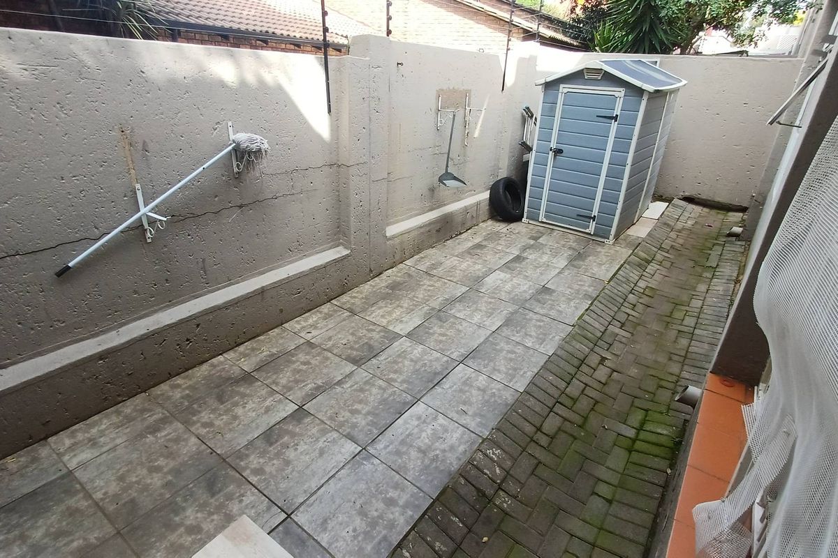 A narrow paved courtyard area featuring a high boundary wall with security fencing and a small plastic storage shed.