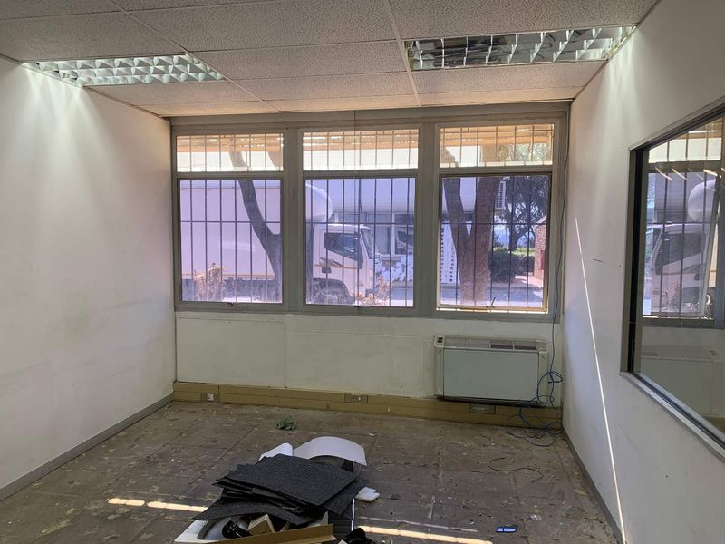 An empty office space featuring a suspended ceiling, security-barred windows, and a wall-mounted air conditioning unit, requiring floor and wall refurbishment.