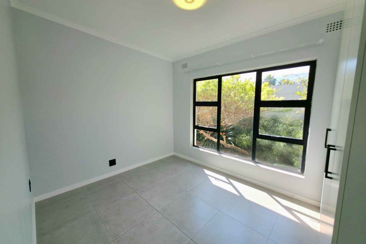 A bright, empty bedroom featuring large black-framed windows, modern grey floor tiles, and a neutral color palette.