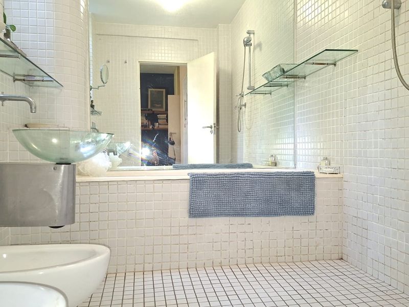 A dated bathroom featuring white mosaic tiling, a built-in bathtub, a glass vessel sink, and a wall-mounted bidet with glass shelving.