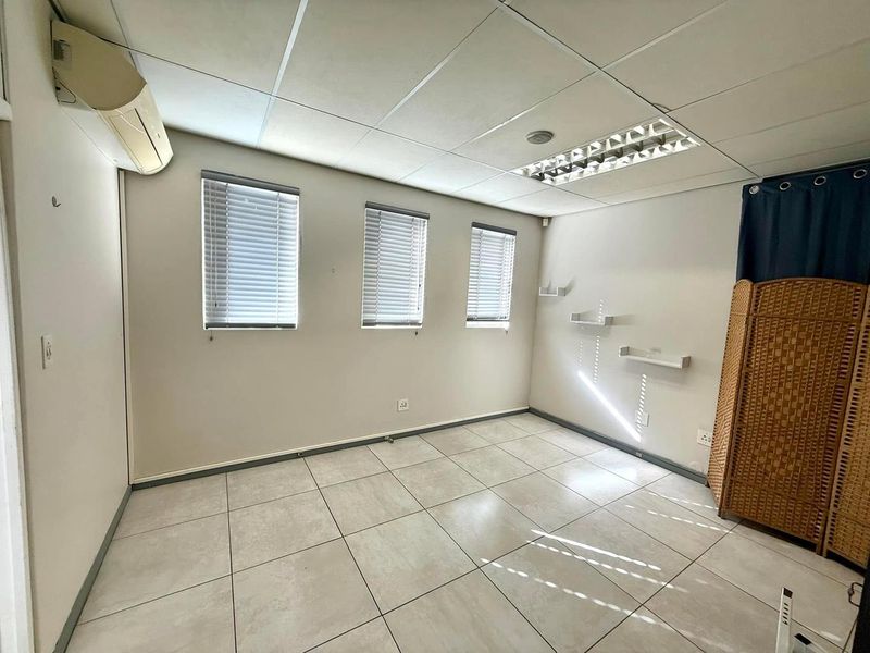 A functional office space featuring tiled flooring, a suspended ceiling with recessed lighting, split-system air conditioning, and natural light through venetian-blinded windows.
