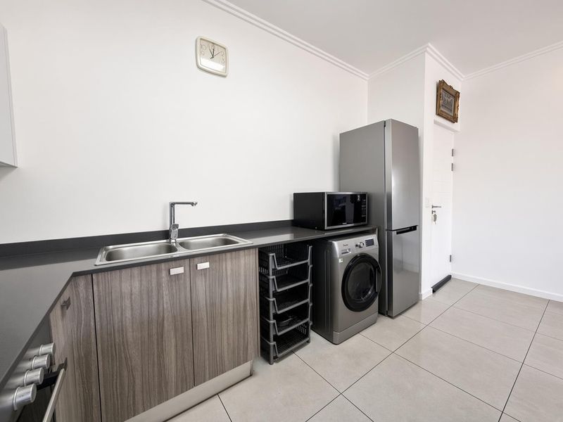 A functional laundry area featuring modern cabinetry, a double sink, stone countertops, and tiled flooring with space for a washing machine.