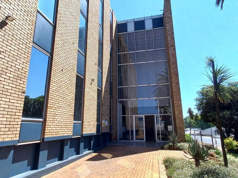 Front exterior view of a multi-story office building featuring a brick facade, large glass curtain wall entrance, and paved pedestrian walkway.