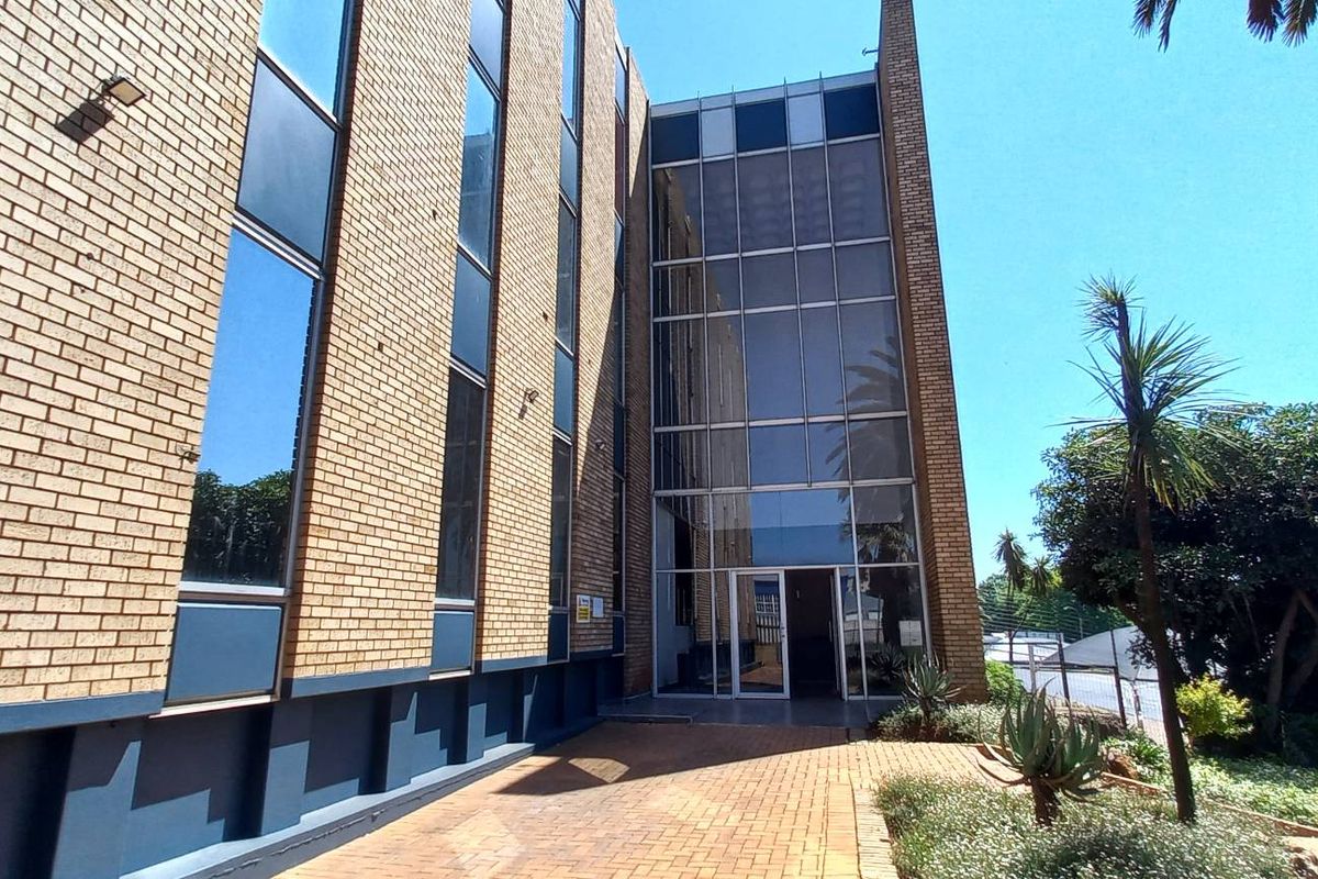 Front exterior view of a multi-story office building featuring a brick facade, large glass curtain wall entrance, and paved pedestrian walkway.