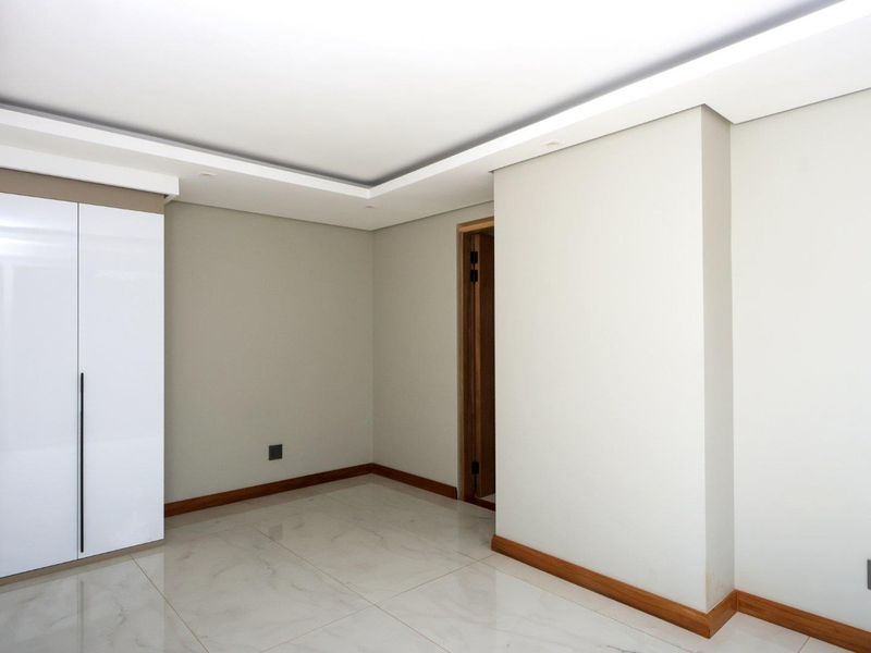 A bright, modern bedroom featuring high-gloss built-in wardrobes, polished tiled flooring, and recessed ceiling lighting with clean, neutral finishes.