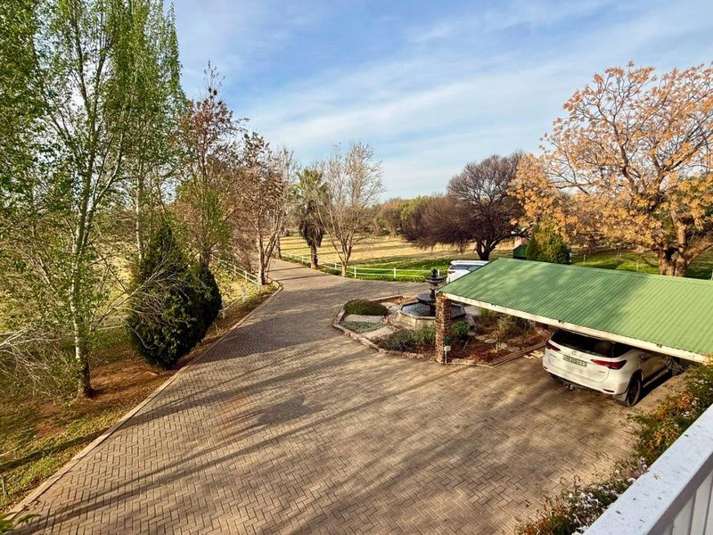 Elevated view of a paved driveway leading to a carport, featuring a central water fountain and established garden surroundings on a small holding.