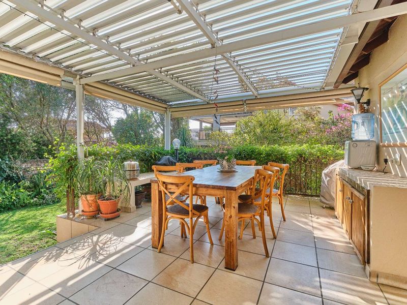 A covered outdoor patio featuring tiled flooring, a corrugated roof, and built-in cabinetry, overlooking a lush garden area.