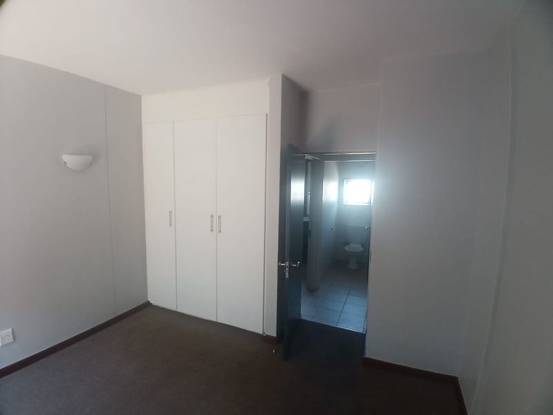 A clean, empty bedroom featuring built-in cupboards, neutral wall finishes, and carpeted flooring, with an adjoining bathroom visible through the doorway.