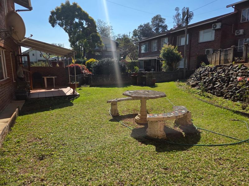 Rear garden area featuring a covered patio with a built-in brick braai, lawn space, and a stone retaining wall.
