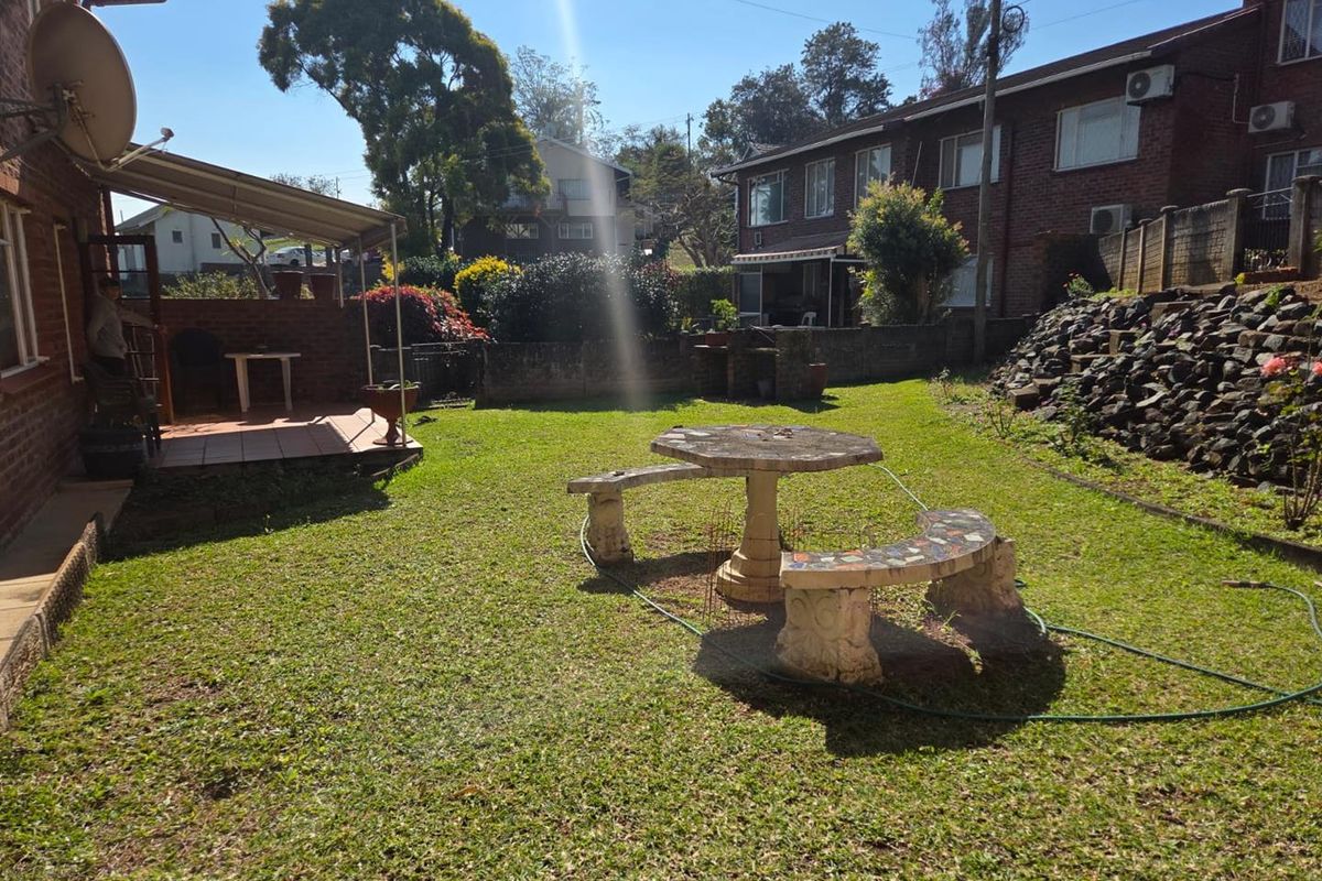 Rear garden area featuring a covered patio with a built-in brick braai, lawn space, and a stone retaining wall.