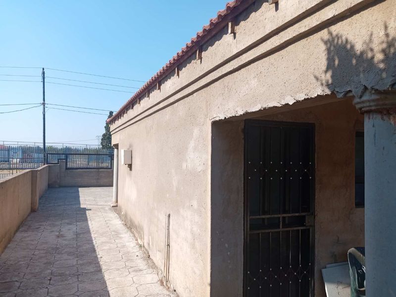 Rear exterior view of the property featuring a paved walkway, security gate, and boundary wall with visible wear to the exterior plaster finish.