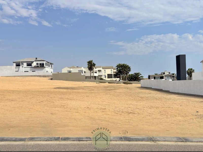 A vacant residential plot of land situated in a developed neighborhood, featuring a level sandy surface and boundary walling.