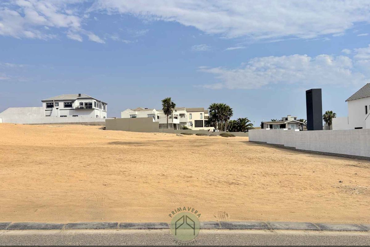 A vacant residential plot of land situated in a developed neighborhood, featuring a level sandy surface and boundary walling.