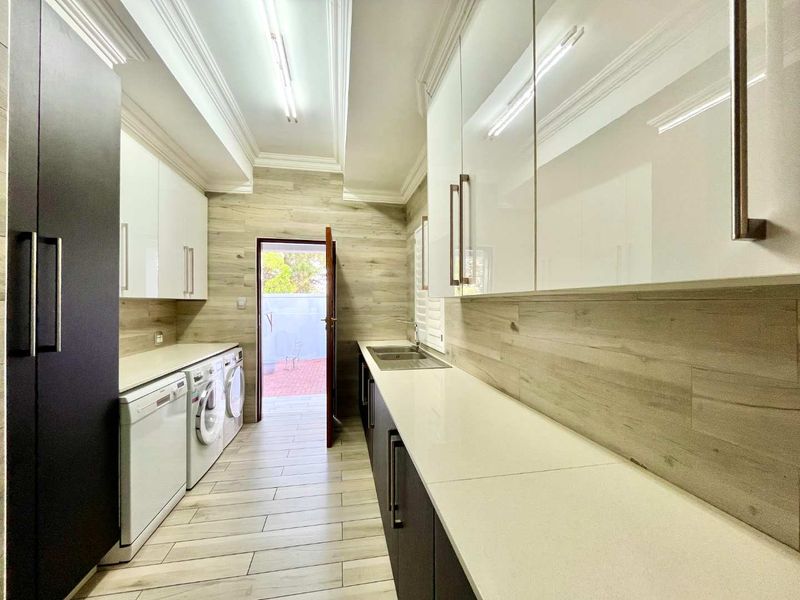 Modern, well-appointed laundry room featuring sleek white and dark cabinetry, stone countertops, double sink, and integrated appliances with wood-look tile flooring.