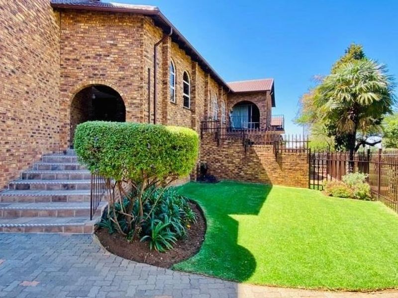 A well-maintained rear exterior view featuring a face brick facade, arched entryway, tiled staircase, and a neat lawn with perimeter metal fencing.