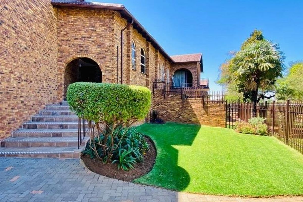 A well-maintained rear exterior view featuring a face brick facade, arched entryway, tiled staircase, and a neat lawn with perimeter metal fencing.