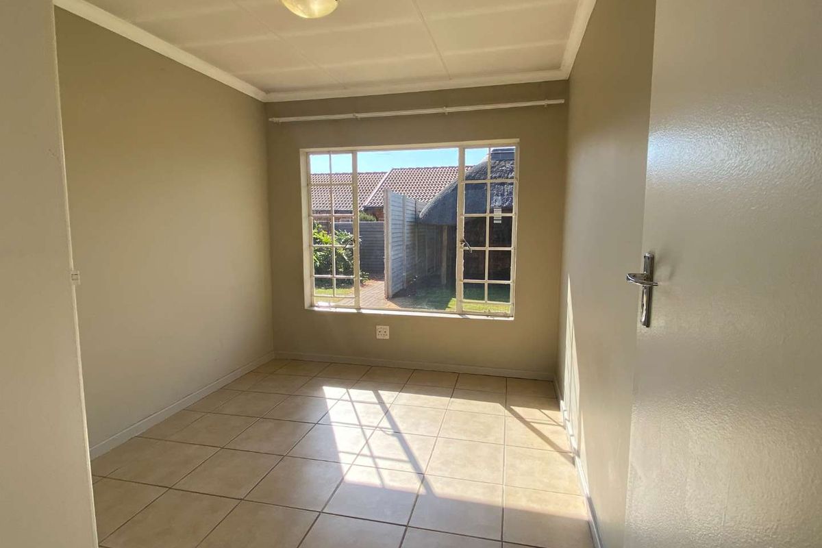 A bright, empty bedroom featuring neutral wall paint, tiled flooring, and a large steel-framed window overlooking the rear garden area.