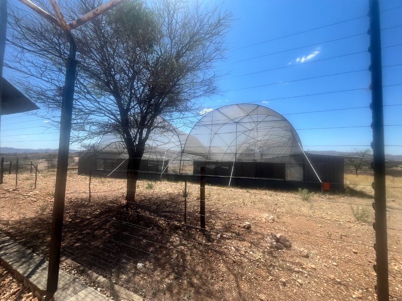 A view of a large outdoor mesh enclosure structure set within a dry, rural landscape, featuring metal framing and perimeter security fencing.
