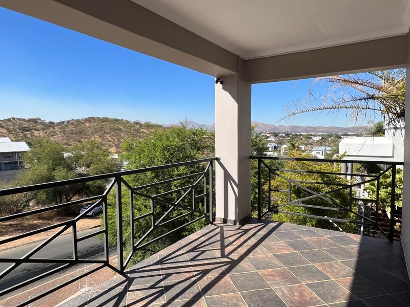 A spacious covered balcony featuring tiled flooring and a secure metal balustrade, offering expansive elevated views of the surrounding landscape and neighborhood.