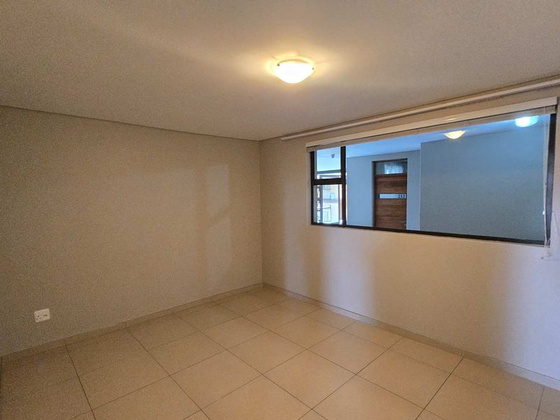 A clean, empty bedroom featuring neutral tiled flooring, a ceiling light fixture, and an internal window looking out toward the apartment entrance.