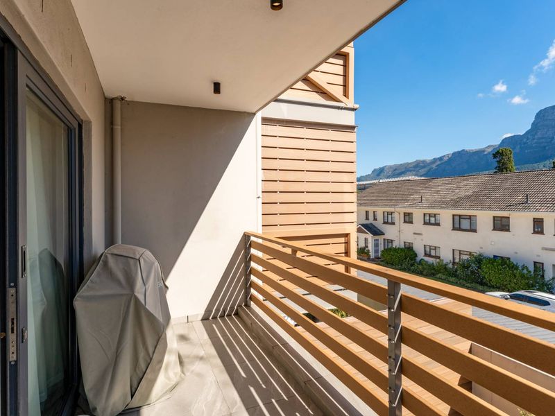 A modern, covered balcony featuring tiled flooring, a timber-look slatted balustrade, and sliding glass door access, offering pleasant neighbourhood and mountain views.