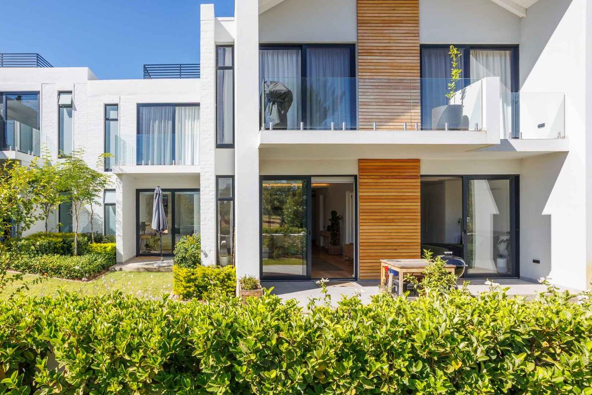 Modern apartment exterior featuring a ground floor patio, upper level balcony with glass balustrades, timber cladding accents, and large sliding glass doors.