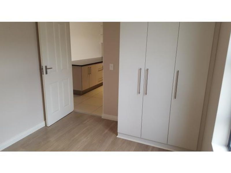 A clean, empty bedroom featuring modern built-in cupboards and light-toned laminate flooring, with a view through to an adjacent kitchen area.