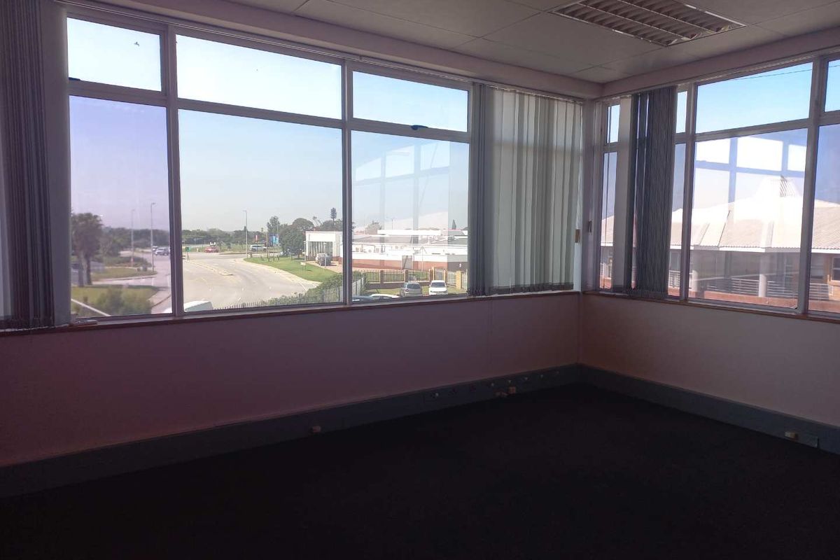 An empty office space featuring large windows with vertical blinds, recessed ceiling lighting, and carpeted flooring, offering ample natural light and exterior views.