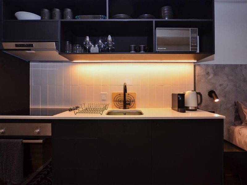 Modern compact kitchen featuring sleek black cabinetry, integrated appliances, tiled backsplash, and warm under-cabinet lighting, seamlessly integrated into the living space.