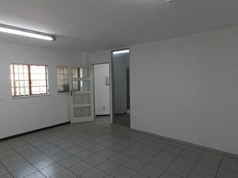 A spacious, empty industrial office area featuring tiled flooring, fluorescent lighting, and security bars on the windows.