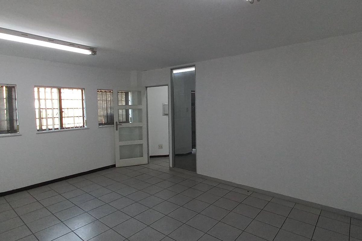 A spacious, empty industrial office area featuring tiled flooring, fluorescent lighting, and security bars on the windows.