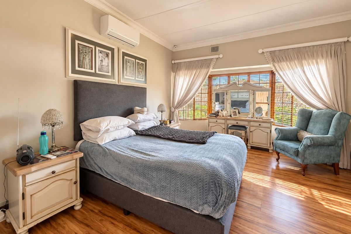 A well-maintained bedroom featuring laminate flooring, a wall-mounted air conditioning unit, and large windows with timber venetian blinds and elegant curtains.
