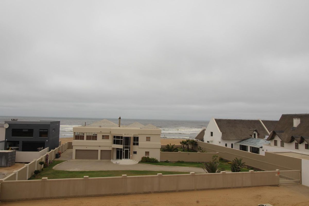 Elevated aerial view of a coastal property featuring a multi-level residence with a paved driveway, perimeter boundary walls, and direct proximity to the beach.