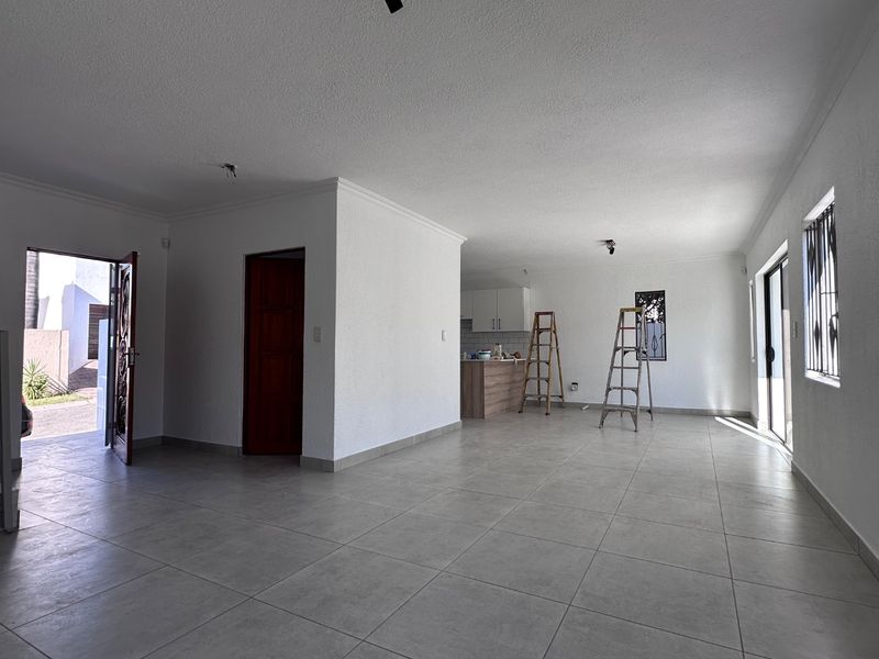 Spacious open-plan living area featuring neutral tiled flooring, integrated kitchen cabinetry, and direct access to the exterior through a security-fitted entrance.
