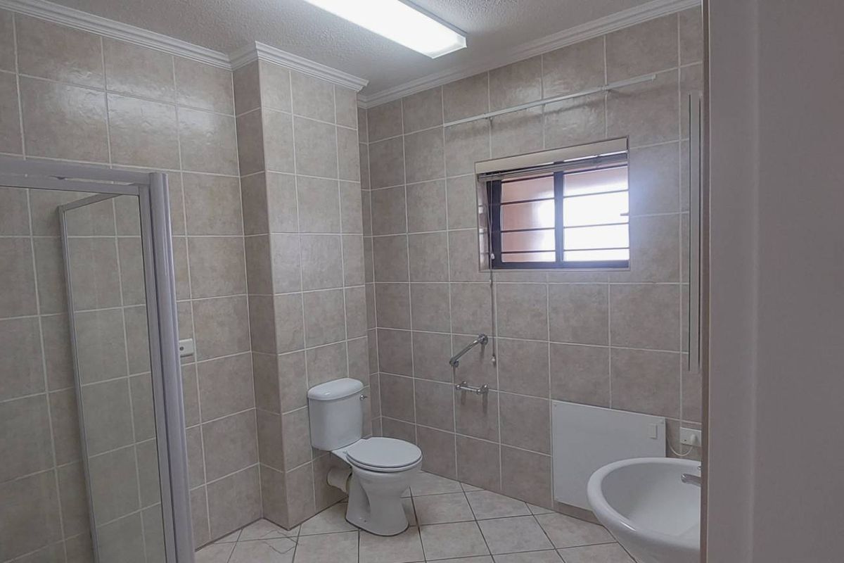 A functional bathroom featuring a shower enclosure, toilet, pedestal basin, and safety grab rails, finished with neutral wall and floor tiling.