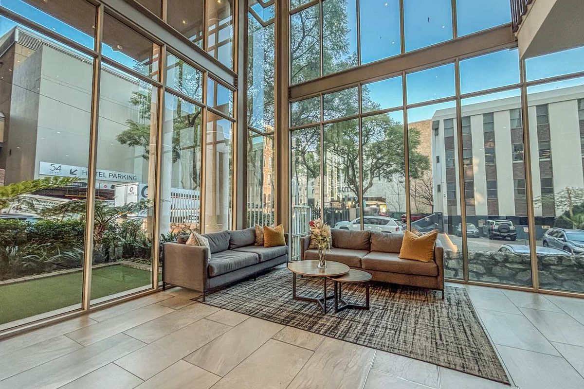 A bright, double-volume living area featuring expansive floor-to-ceiling glass windows that provide abundant natural light and views of the surrounding urban environment.